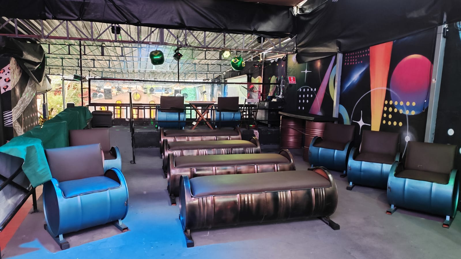 Lounge da Villa com puffs de barril e mural colorido — assinatura visual da casa